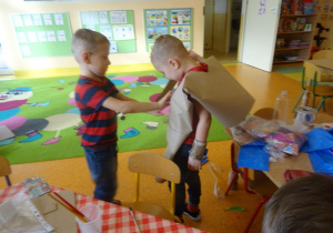 Jeden z chłopców ma założony duży szary arkusz papieru, na który drugi chłopiec nakleja elementy dekoracyjne.